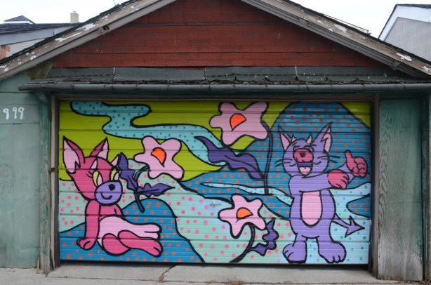 mural with two cartoon like animals, one pink and the other blue, beside a blue mountain along with some pink flowers, a mural on a garage door 