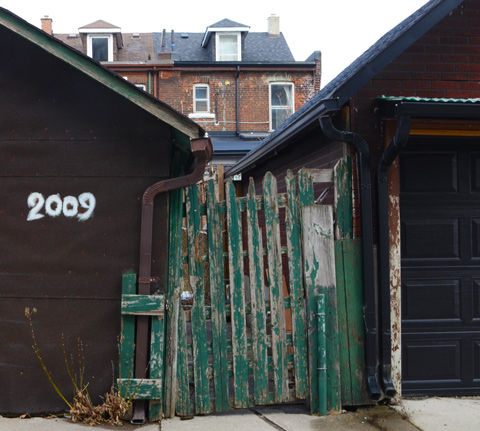 in an alley, two old garages with a crooked gate with peeling dark green paint, house in the background