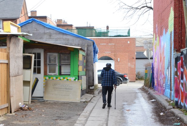 an older man with a cane and a plaid jacket walks down an alley, back to the camera