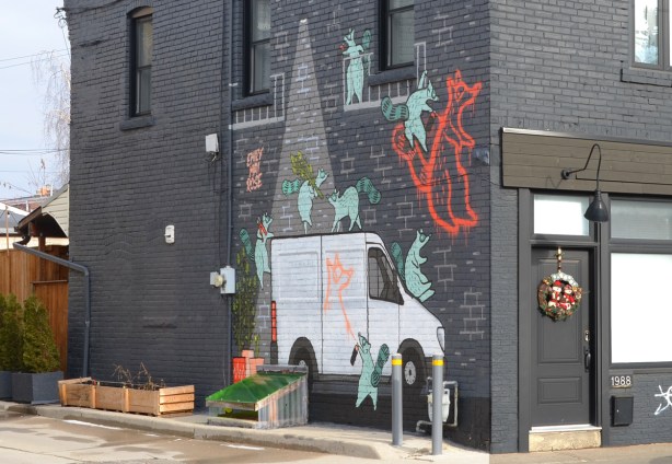 emily may rose mural on the side of a dark grey building, raccoons playing around a white cube van, with orange spray paint