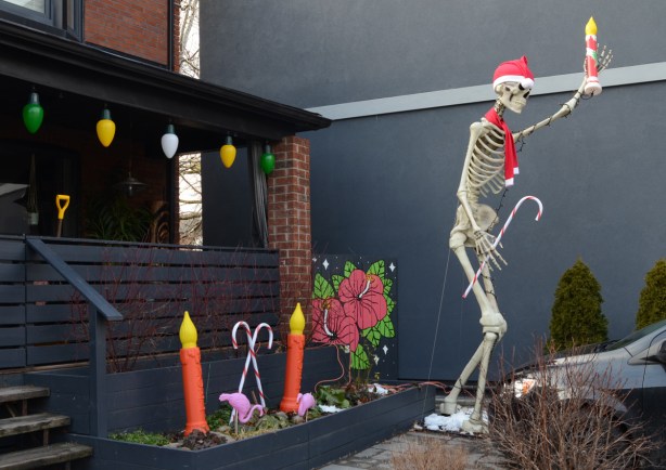 large skeleton outside in front yard with Santa hat and red scarf, holding a plastic candle light, other Christmas decorations on porch of house