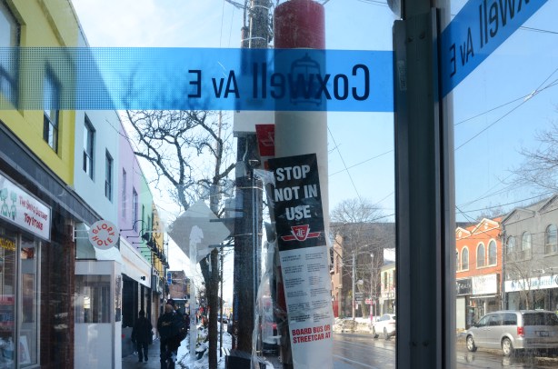 looking through the clear side of a bus shelter at Coxwell Ave., looking west along Gerrard St.