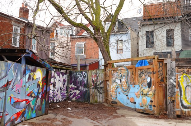 a dead end in an alley where all the fences and gates are covered with murals, a large tree, the backs of two storey houses in different materials and colours, brick, wood, 