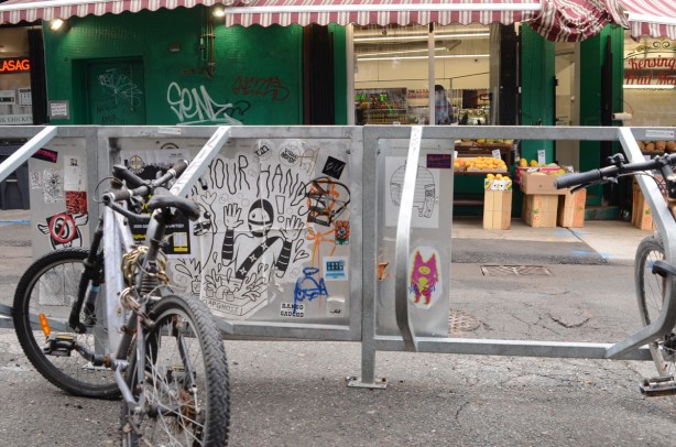 bke parked at bicycle stand with graffiti slaps on it, across street from fruit and vegetable market with green walls and red and white striped awning, Kensington market area of Toronto