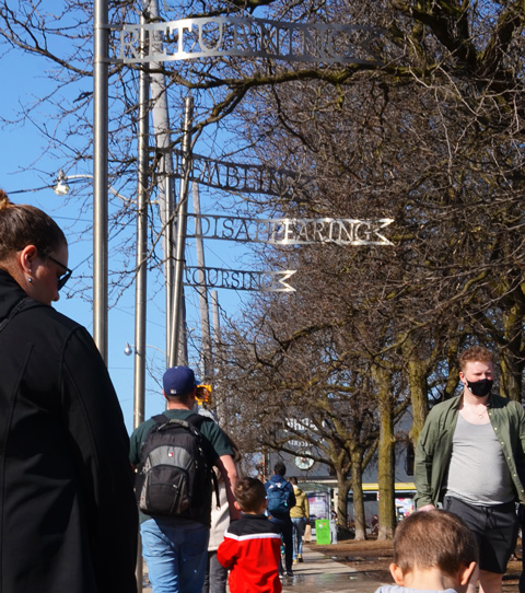 people walking on sidewalk, walking past 4 metal poles holding up words in metal