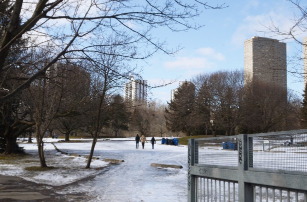 three people walking across a snow covered park towards highrise buildings