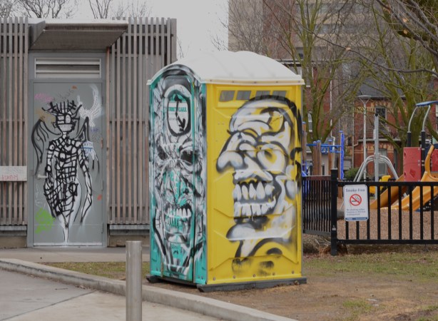 a blue and yellow porta potty covered in macabre street art, in bellevue park