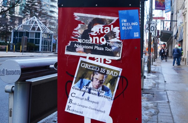 graffiti and posters on the side of a red metal newspaper box on the sidewalk. I'm feeling blue sticker, and an anti development poster saying greed is bad 