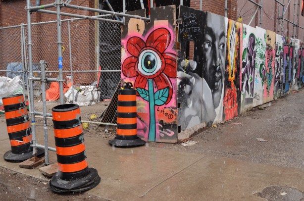 black and orange traffic pylons beside the entrance to Graffiti Alley with a red one eyed daisy painting by life co beside it, then a row of paintings on hoardings