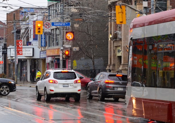 TTC streetcar westbound on Queen West approaching Bathurst, snow falling, other cars waiting for red light, 