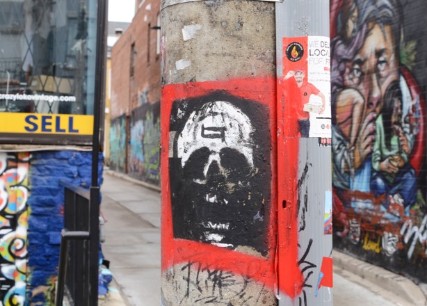 poster graffiti of a white skull on black background, large red border, on a pole, with alley street art in the background