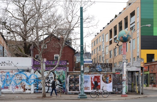 man on sidewalk on Spadina, walking past hoardings with graffiti and street art and adverts, one mural is a large skull with outreached bony arms, 