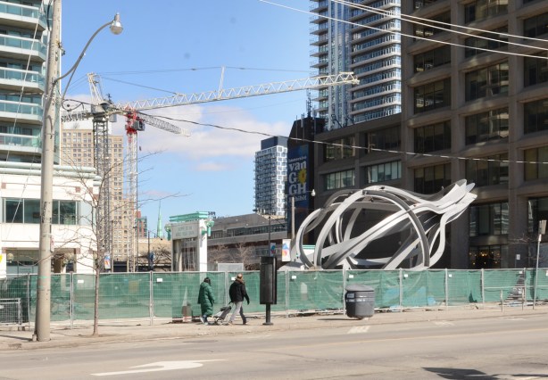 two people walking past a site with a green fence, a sculpture behind the fence, new buildings and new construction in the background