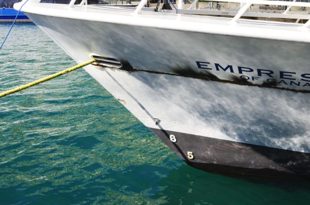 front end of boat, Empress of Canada, white and black hull, dirty, tied to pier with yellow rope, reflections of it in the water of the harbour