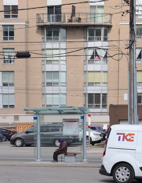 man sitting in a bus shelter, brick apartment building behind him