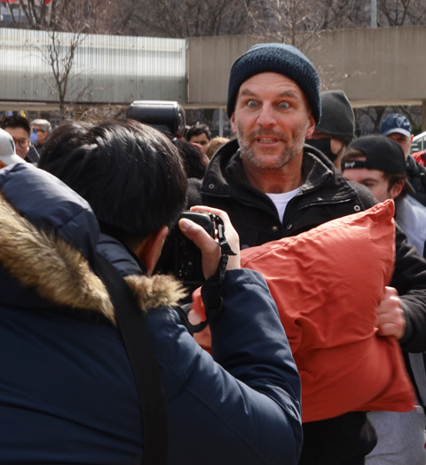 people participating in pillow at nathan phillips square, outdoors, fighting with pillows, man with his toque over his eyes, a man with orange pillow case making faces at another person taking his picture