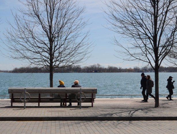 two people sitting on a bench, one with yellow toque and the other with yellow turban, other people walking past, on the waterfront