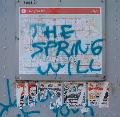 graffiti text sprayed on map and wall of bus shelter says the spring will (illegible), partially removed posters below that for communist party 