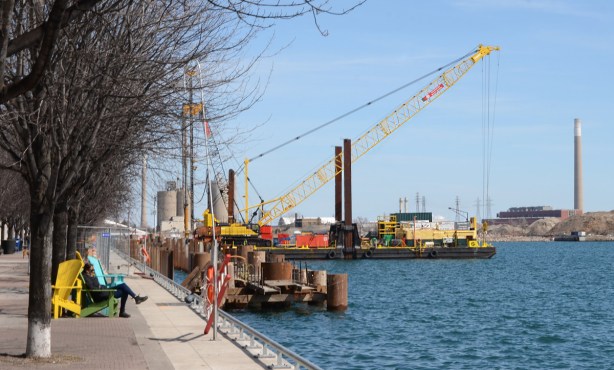 a person sitting in a muskoka chair on the waterfront near a water work site with barge, and rusty metal pylons in the water, port lands in the distance