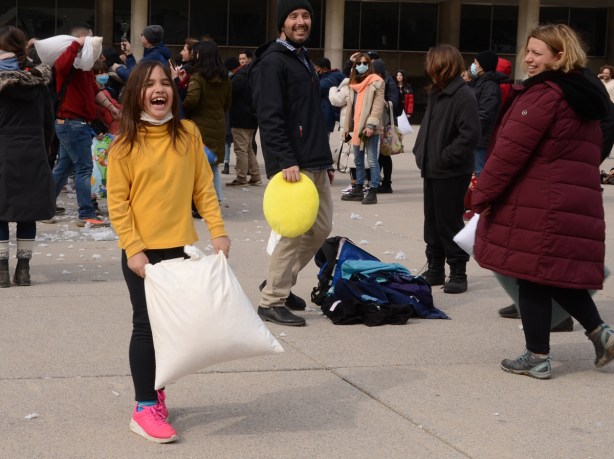 people participating in pillow at nathan phillips square, young girl in yellow top and pink shoes is laughing, she is holding a pillow