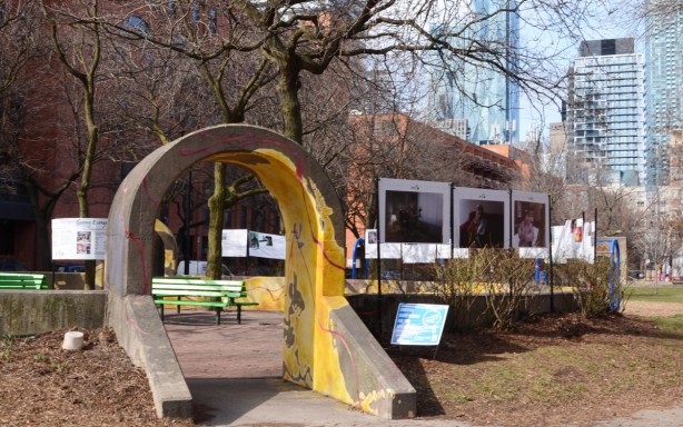 small yellow concrete arch leading to a circular garden in a park, with posters as part of an art exhibit