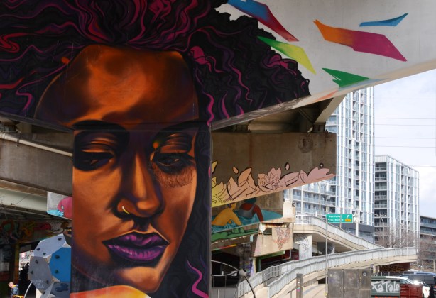painted concrete pillar in Underpass park of a woman with purple lips, city in the background - sidewalk ramp up to Queen Street, condo