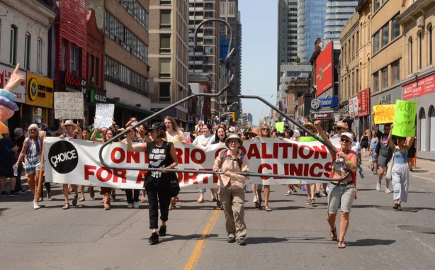 giant coat hanger being carried in front of banner for ontario coalition for abortion clinics, dyke march