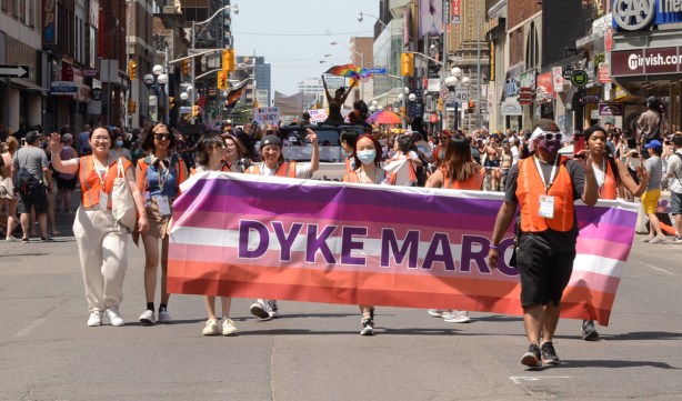 dyke march banner at the beginning of a parade, purple and orange stripes, carried by 5 women, on Yonge Street