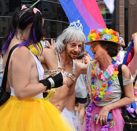 man in grey hair, silver crown, bending over to talk closely to woman in hat covered with multi coloured flowers, start of pride parade