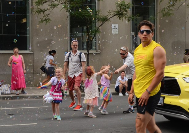 kids playing in bubbles while a man in a yellow t shirt walks past, on a street,