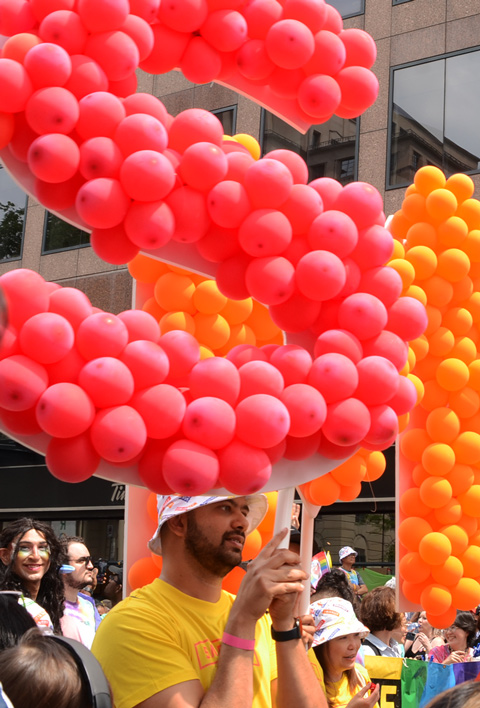 large letters S and I made of balloons in a pride parade, held by a man in yellow with white hat