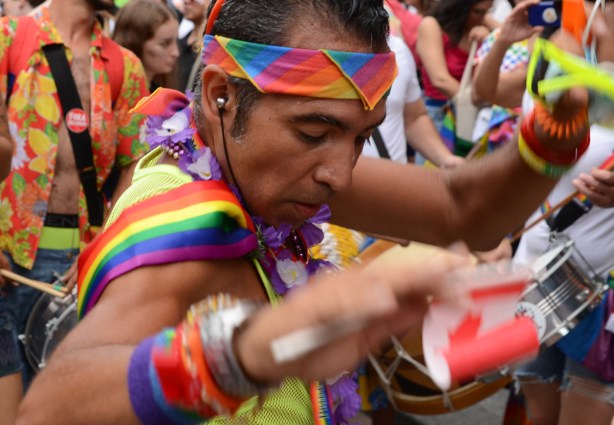 man with rainbow bandana around head, and scarf on back as well as many bracelets, holding small Canadian flag, dances in the street for pride parade