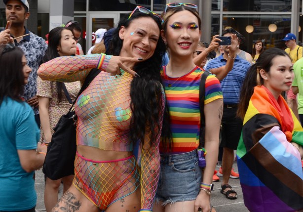 two people at pride parade, one in rainbow striped shirt and the other in rainbow netting outfit with stickers over nipples, thong underwear under neath