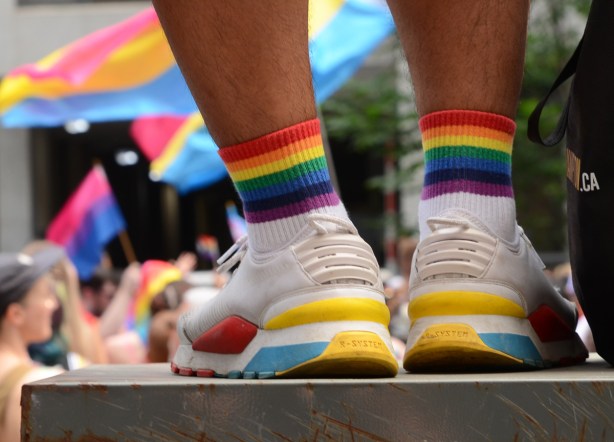 rainbow striped ankle socks, and running shoes with red, yellow, and blue on them, standing on a sidewalk box to get a better look at passing pride parade