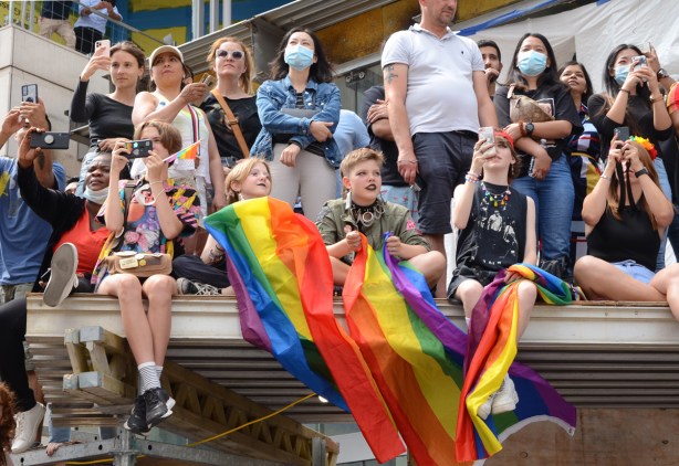 people sitting on scaffolding over sidewalk by construction site to watch pride parade, two have rainbow flags 