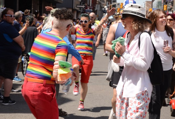 two women in matching red pants and rainbow striped tops dancing in parade, and laughing with other people