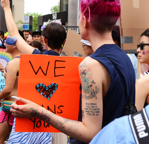 Toronto Pride Parade, person with tattoo on upper arm holding an orange sign that says we love trans youth. Tattoo says In a dream you saw a way to survive and you were full of joy 