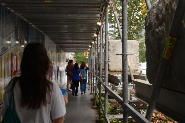 a young woman with long hair holds a cup of coffee as she walks on sidewalk under protective covering beside construction at Yonge and Bloor