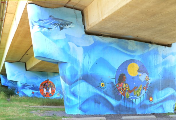 subway tracks cross above the Humber River at Old Mill station. The concrete pillars that support the subway bridge have been covered with many watery blue First Nations themed murals. fish swimming in watery blues around central medallions with images