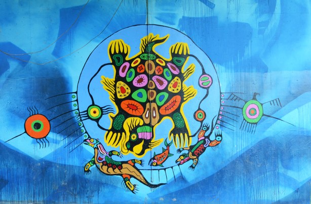 First nations mural on concrete pillars holding up subway bridge over the Humber River, featuring a turtle shaped animal with a bear head, with its mouth open hunting for fish 