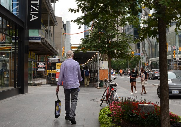 an older man with thinning grey hair walks on sidewalk, Bloor Street, towards Yonge, and scaffolding over sidewalk because of construction