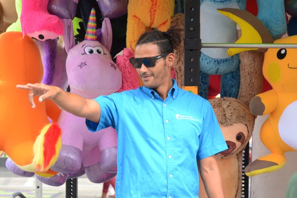 man in dark sunglasses and turquoise shirt, standing in front of wall of giant stuffies, pointing at someone