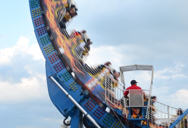 a man in a red t shirt sits in a booth as he controls a ride at the Canadian National Exhibition, a ring with riders whizzing past