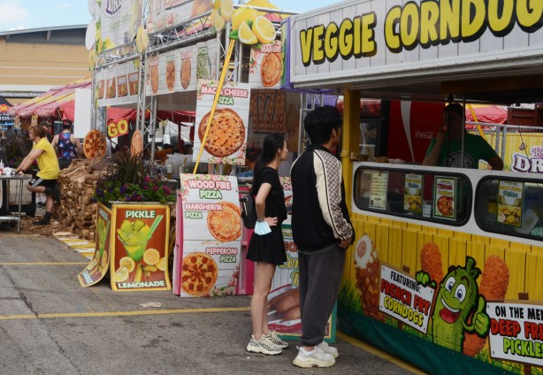 two people waiting at a veggie corndog vendor, the vendor next to them is advertising pickle lemonade 