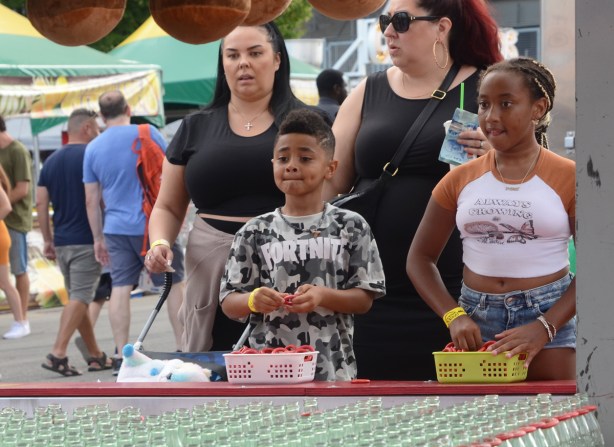 two kids and two women at a ring toss game, small red rings over the neck of coke bottles 