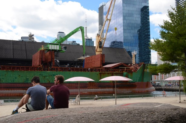 two men sit on the rock at Sugar Beach, with green sugar ship docked at Redpath sugar, city skyline behind