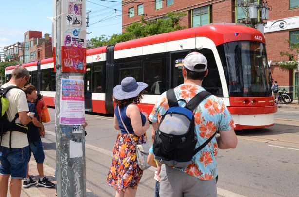 hot summer day, people waiting for an arriving TTC streetcar on College Street, woman in a wide brim blue hat and flowery skirt, man in blue Hawaiian shirt with orange and yellow flowers