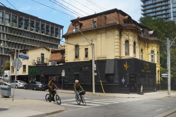 corner of Dundas and Mutual, two men on bikes, older house on corner with yellowish brick and mansord roof, now a cannabis shop