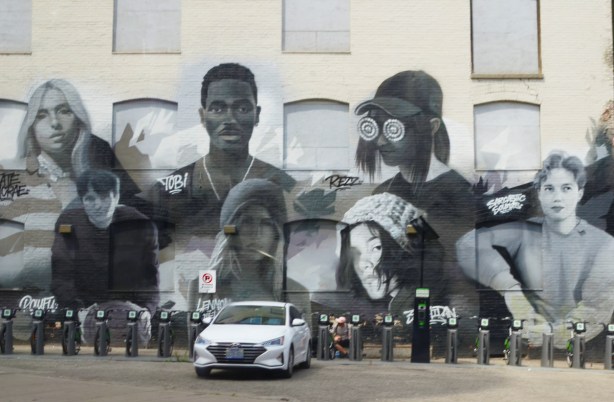 large black and white mural on Dundas Street, white car parked in front of it. Mural features portraits of people 