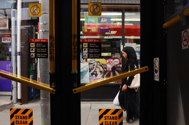 a young woman in black hajib and white bag walks past closed doors of a bus